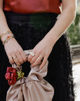 Person holding a beige clutch with floral details, wearing a black skirt, vintage watch band bracelets and a silver and quartz statement cocktail ring.