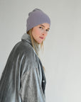 Woman wearing a gray coat and light purple 8.6.4 cashmere beanie against a plain background