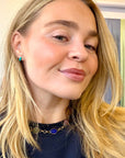 Smiling woman in a navy top wearing small turquoise stud earrings and a gold necklace with blue stones