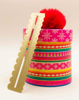 Brass wavy ruler with a scalloped edge leaning on a pink holiday gift box topped with a red pom-pom