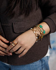 Close up of person's wrist and hand wearing 5 vintage gold-filled bracelets including tiger's eye, opal, carved peking glass, jade and two, gold rings with hand-painted glass centers.