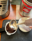 Handmade heart-shaped metal coffee scoop measuring ground espresso beside an espresso machine