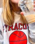 Close up of woman wearing pearl and leather cord necklace over a Syracuse basketball sweatshirt drinking iced coffee