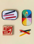 Open Block Design tin with crayons beside a colorful closed tin and its Top Draw sleeve