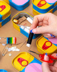 Hands threading a needle over an open Sew Cute sewing kit tin with pins and colored thread