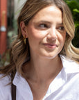 A close-up shot of a woman with blonde-highlighted hair wearing a white button-down shirt and a gold dangle earring, set against a blurred outdoor background.