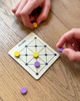 Hands playing Picaria with yellow and purple tokens on a square wooden board
