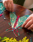Hands threading a hand-printed paper moth in teal, red and gold onto the garland string