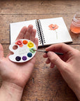 Hand holding a mini paper watercolor palette while painting a poppy in a sketchbook