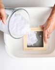 Hands pouring paper pulp into a wood mold set in a tub of water