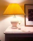 Small glass hourglass on a pink nightstand next to a lamp with a yellow pleated shade