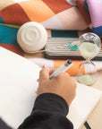 Hand writing in a notebook beside a glass hourglass and a striped pencil case