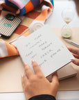Hand holding the white Poète notebook on a desk with an hourglass and phone
