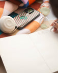Poète notebook lying fully flat while someone writes next to a sand timer
