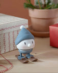 Blue-capped Hoptimist skier on a wooden table beside wrapped gifts and a potted fir