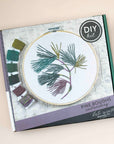 Kate Wyatt Pine Boughs embroidery kit box with a stitched pine branch in a wooden hoop