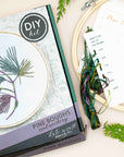 Pine Boughs embroidery kit box beside its stitched hoop and instruction sheet