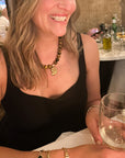 A woman in a black sleeveless top smiling while wearing a gold pendant necklace and bracelets, holding a clear wine glass in a restaurant setting.