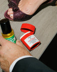 Man's hand holding a bottle of Man Oil next to an opened stick of Ouch Balm next to a woman's foot in a jeweled, burgundy sandal.