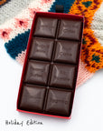 Close-up of an opened Miss Maude’s Holiday Edition dark chocolate bar with eight flavored squares displayed on a patterned sweater background.