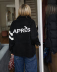 Back of a woman wearing Pink Pineapple Après ski black varsity cardigan with white stripes on one sleeve and Après graphic text on the back.