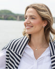 A smiling blonde woman in a white button-down shirt and a black and white striped knit cardigan, wearing a delicate gold necklace and earring, photographed outdoors with a blurred body of water and green hills in the background.