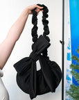 Black bow crossbody bag with ruched strap, shown close up against a white wall.
