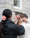 Two young women in pontyails adjusting pink satin rosette scrunchies in front of a stone building.