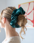 Close up of the back of a woman with blonde hair wearing a giant, oversized satin midnight teal green scrunchie over a messy bun.