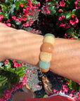 A person's wrist wearing a beaded stretch bracelet made of smooth, rounded stones in muted shades of green, peach, cream, and light blue, photographed outdoors against a background of pink flowers and green foliage.