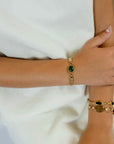 A close-up of a person's wrist wearing a gold-toned watchband bracelet with a prominent jade stone, paired with layered gold bracelets on another wrist, set against a soft white top.