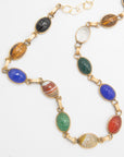 A gold-tone chain necklace featuring multiple oval cabochon gemstones in various colors including blue, green, red, brown, black, and white, arranged in a pattern on a white background.
