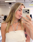 A woman taking a mirror selfie in a boutique, wearing a cream-colored ribbed tube top and a gray silk tie-necklace. She is holding a light beige smartphone.