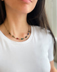 A close-up of a woman wearing a delicate gold chain necklace with small multicolored scarab gemstones, paired with a white crew-neck t-shirt.
