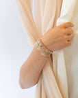 Woman wearing a stack of gemstone beaded bracelets on her wrist while holding sheer peach and cream fabric drapes.