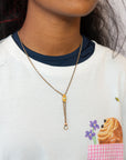 A close-up of a person wearing a delicate gold Victorian-era chatelaine slider necklace with a small gold charm and drop ring, styled over a white ringer T-shirt with a navy collar and a cartoon pastry pocket.