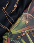 Gold-filled Victorian-era chatelaine necklaces with dog clip clasp connectors and small floral charms, arranged on a dark navy fabric and a tropical leaf print surface.