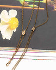 An antique bronze double-strand Victorian-era gold-filled chatelaine necklace with a small diamond-shaped crystal pendant and a larger square crystal pendant, displayed on a dark book cover next to a white flower.