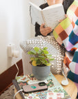 A cozy lifestyle scene showing a person wrapped in a colorful plaid blanket and reading a notebook on a wooden chair. In the foreground, a small potted succulent sits on a wooden table next to a striped phone case and a small hourglass.