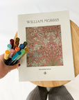 Hand holding a bundle of Tombow dual brush pens beside a William Morris coloring book on a wooden stool.