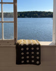 A dark olive green towel with white polka dots draped over a windowsill, with a scenic view of a blue lake and tree-lined shore in the background.