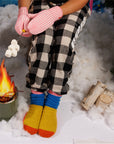 Person pretending to roast marshmallows over a fake fire wearing Verloop striped mittens, colorful house-knit socks and checkered pants.