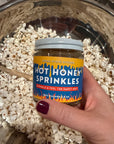 Hand holding a jar of Wood Stove Kitchen Hot Honey Sprinkles over a bowl of popcorn