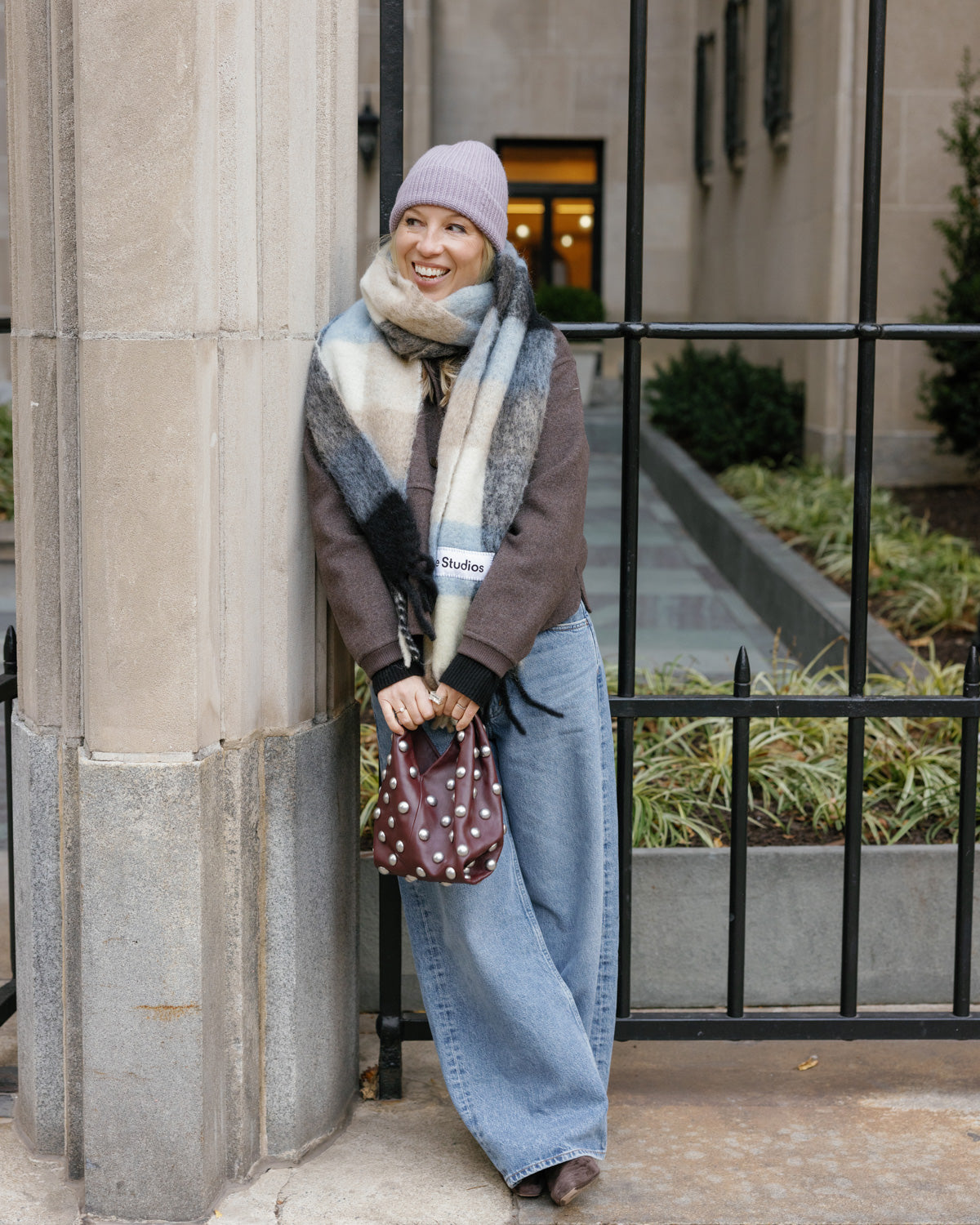 Woman wearing a plaid scarf, brown coat, blue jeans and 8.6.4 cashmere knit beanie in light purple, standing against a building exterior.