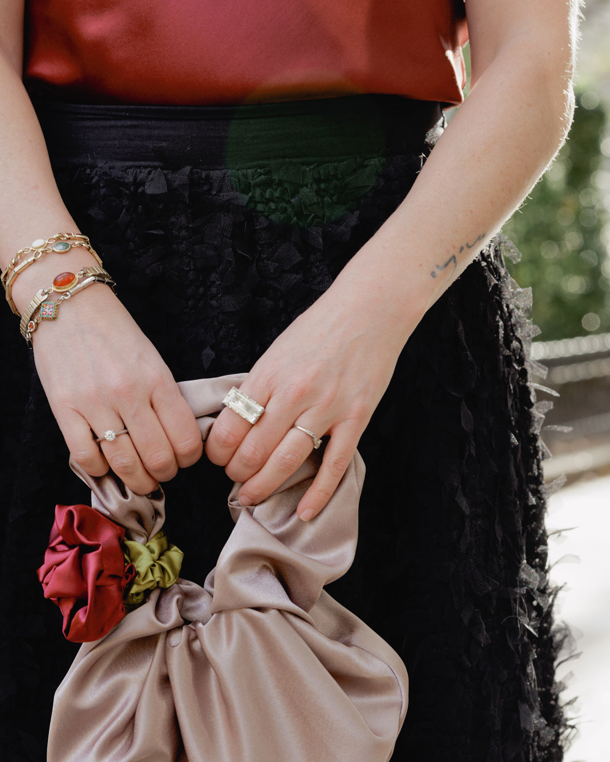 Person holding a beige clutch with floral details, wearing a black skirt, vintage watch band bracelets and a silver and quartz statement cocktail ring.