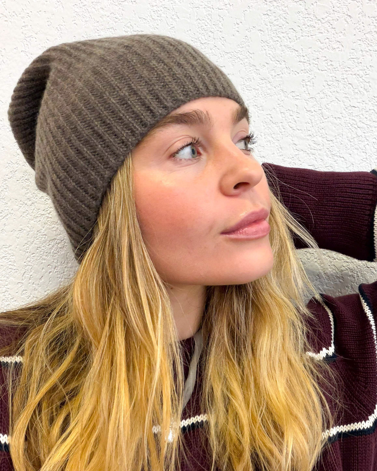 A woman with blonde hair wearing a ribbed brown cashmere knit beanie hat and a striped burgundy sweater, looking to the side against a textured white wall.
