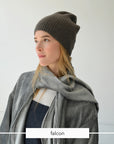 A woman wearing a dark brown ribbed knit beanie, a gray scarf, and a navy top, shown in profile against a light gray background. A white label at the bottom reads 'falcon'.