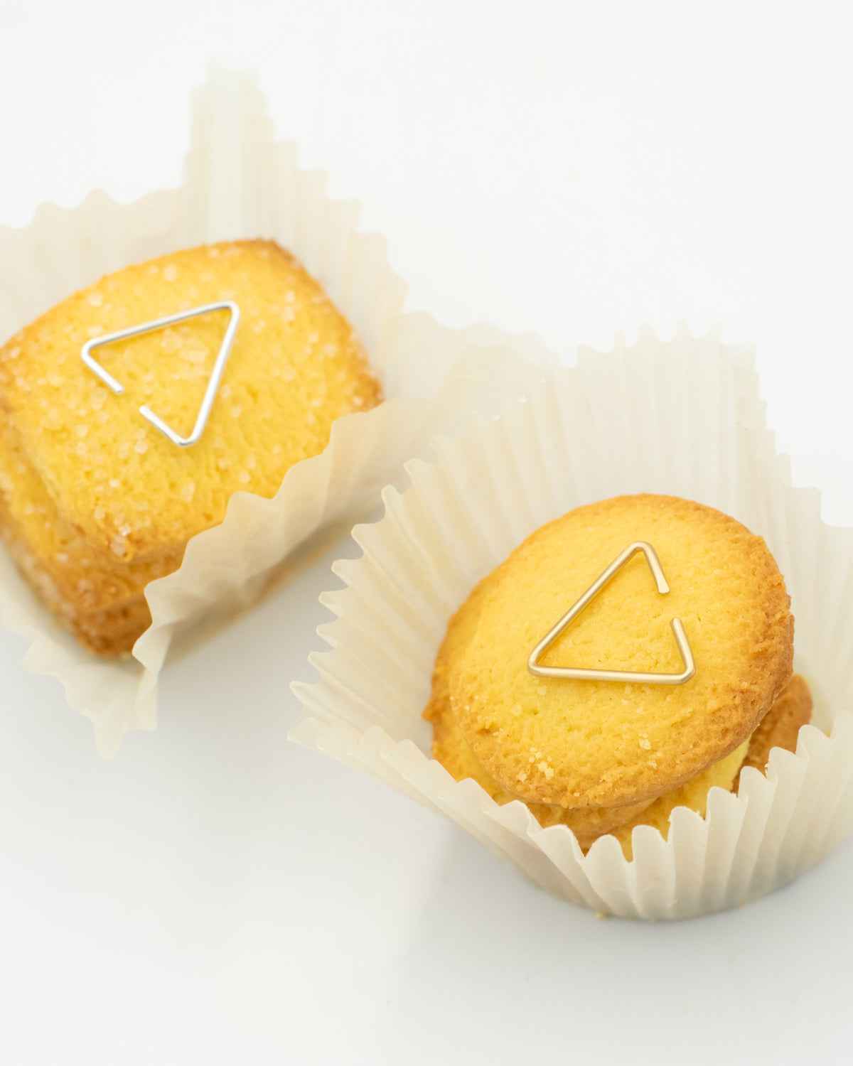 Yellow cookies in paper liners with two 8.6.4 triangle ear cuffs, one brass and one silver, on a white background.