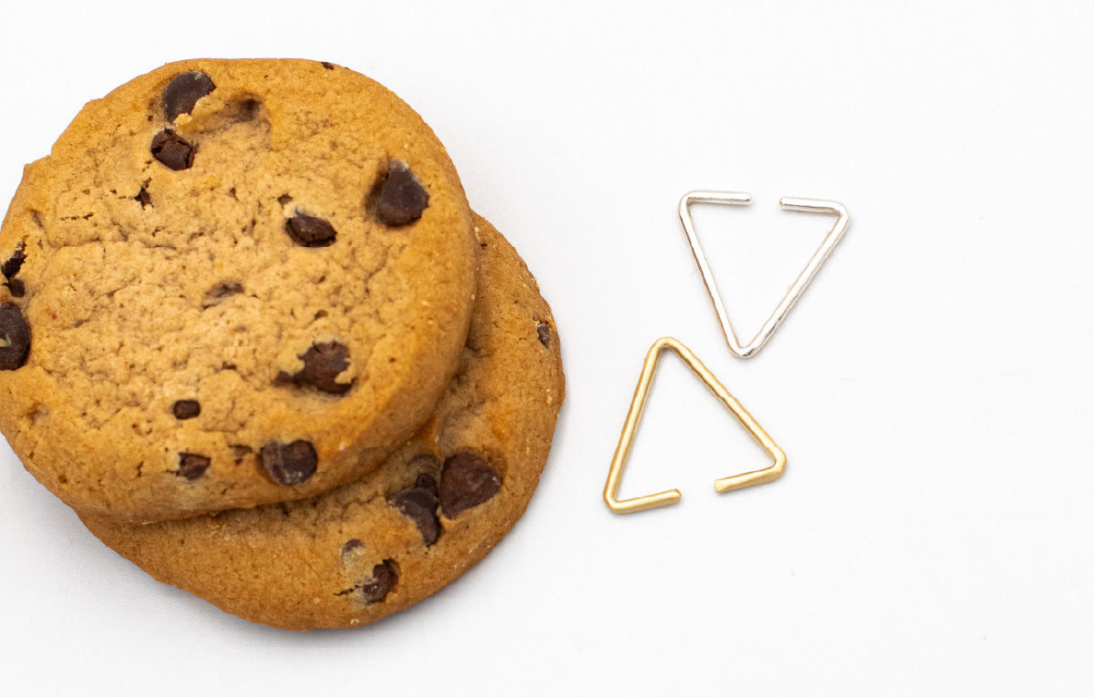 Two 8.64 triangle ear cuffs: one brass, one silver, on a white table next to chocolate chip cookies.