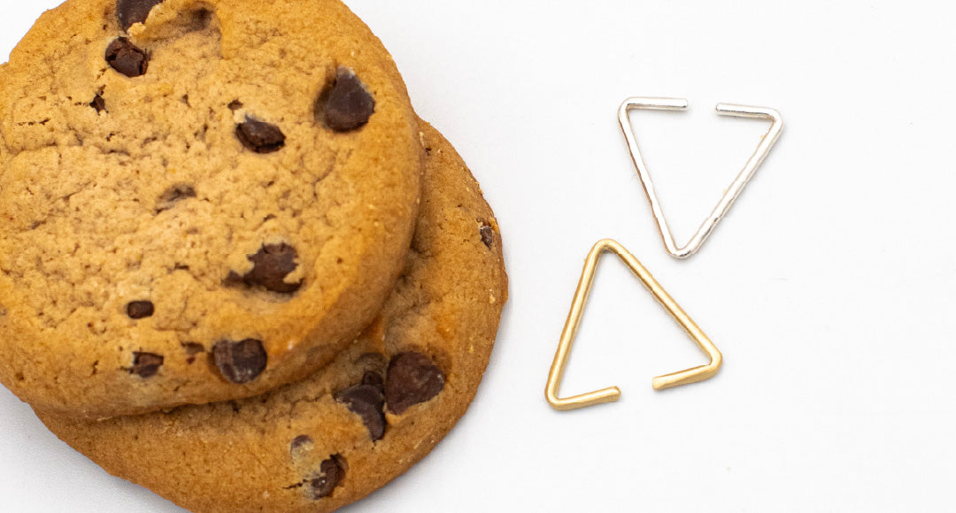 Two 8.64 triangle ear cuffs: one brass, one silver, on a white table next to chocolate chip cookies.